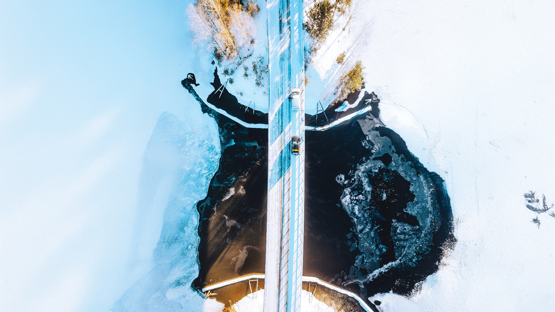 Luchtfoto van een besneeuwde brug met twee auto’s die deelnemen aan de Ice Road Rally, rijdend over een bevroren rivier met donkere wakken, omringd door sneeuw en ijs in een winters landschap.
